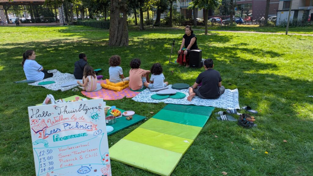 Een foto van een groep kinderen in een park. De begeleider leest aan hun voor