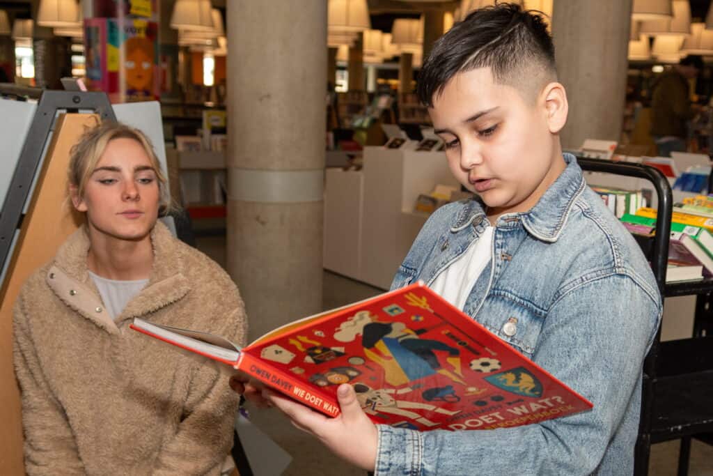 Een foto van een kind dat een boek voorleest aan zijn begeleider in de bibliotheek